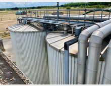 Industrieanlage mit metallischen Silotanks und Rohrleitungen, umgeben von einer ländlichen Umgebung mit Feldern und Bäumen im Hintergrund. Ein erhöhtes Geländer führt über die Tanks, während die Landschaft Windkraftanlagen in der Ferne zeigt.