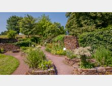Ein Kräutergarten mit geschwungenen Wegen, die von Blumen- und Pflanzenbeeten gesäumt sind. Die Beete sind von niedrigen Steinmauern umrahmt, im Hintergrund steht eine dicht bewachsene Natursteinmauer, und dahinter befinden sich große, grüne Bäume.