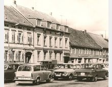 Schwarz-Weiß-Fotografie einer Straße mit geparkten Autos aus der DDR-Zeit vor historischen Gebäudezeilen mit Schindeldächern und Fassaden mit Fensterläden. Im Hintergrund befinden sich Schilder von Geschäften.