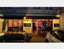 Außenansicht einer Bar oder Kneipe mit dem Namen „Casa“, beleuchtet in warmem Licht. Die Fassade zeigt große Fenster mit Dekormustern, eine niedrige Theke dahinter und Tische im Innen- und Außenbereich, während parkende Autos im Vordergrund zu sehen sind.