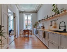 Helle Küche mit Holzboden, langen Arbeitsplatten aus Holz, einer schwarzen Spüle und Wandregalen mit Gewürzdosen. Im Hintergrund befindet sich ein Esstisch mit Stühlen vor einem großen, klassischen Fenster mit Blick auf einen Innenhof.