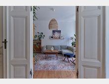 Blick durch eine geöffnete weiße Doppeltür in ein Wohnzimmer mit einem grauen Ecksofa, einem Holztisch auf einem gemusterten Teppich und dekorativen Elementen wie Zimmerpflanzen und Bildern an der Wand. Eine geflochtene Hängeleuchte hängt von der Decke, und der Boden ist aus Holz.