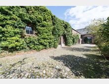 Eine historische Backsteinfassade, teilweise mit dichtem Efeu bewachsen, neben einem gepflasterten Hof. Im Hintergrund ist ein großes Eingangstor zu sehen, das zu einem Gebäude oder einer Halle führt.