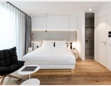 Helles, modernes Hotelzimmer mit einem Doppelbett, einem gepolsterten schwarzen Sessel und einem kleinen weißen Beistelltisch auf Holzboden. Im Hintergrund eine beleuchtete Wand hinter dem Bett, ein Vorhang an der Fensterfront und ein offenes Badezimmer mit einer verglasten Dusche.