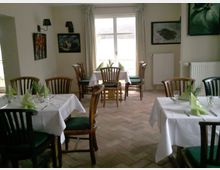 Kleines Restaurant oder Café mit mehreren Holztischen und -stühlen, die mit weißen Tischdecken gedeckt sind. Die Tische sind mit Gläsern, Besteck und grün gefalteten Servietten dekoriert; an den Wänden hängen Bilder, und durch ein großes Fenster fällt Tageslicht in den Raum.