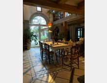 Gemütlicher Essbereich in einem rustikalen Interieur mit Holzbalken, einem großen Tisch umgeben von Holzstühlen und einer klassischen Hängelampe. Im Hintergrund ein großes, gewölbtes Fenster mit Blick auf einen Garten sowie zahlreiche Pflanzen und dekorative Elemente.