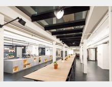 Beleuchteter moderner Gemeinschaftsraum mit einer langen Holztischreihe und schwarzen Stühlen, weißem Pfeilerdesign und einer Küchenzeile mit Regalen und Dekorationen im Hintergrund. Glasdeckenpaneele und industrielle Akzentleuchten ergänzen das offene Ambiente.