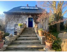 Haus mit Satteldach und blauer Eingangstür, umgeben von Kletterpflanzen an der Fassade. Eine steinerne Treppe führt zum Eingang, gesäumt von Blumentöpfen und kahlen Sträuchern; im Hintergrund Garten mit Bäumen.