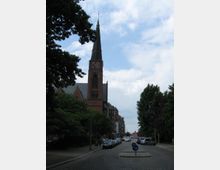 Straßenszene mit einer Backsteinkirche im neugotischen Stil, die einen hohen Turm mit Spitze und Uhr zeigt. Links und rechts der gepflasterten Straße stehen parkende Autos, Bäume und einige Wohngebäude.