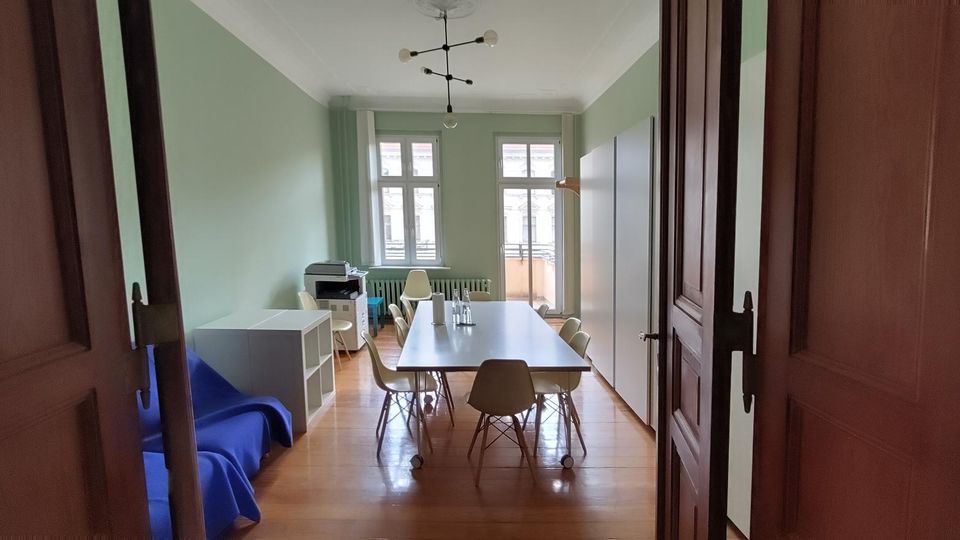 Blick durch eine geöffnete Holztür in einen schmalen, hellen Büroraum mit hohen Decken, mintgrünen Wänden und Holzboden. In der Mitte steht ein langer Besprechungstisch mit Stühlen; am Ende des Raums sind große Fenster mit Heizkörper darunter sowie ein Schrank und ein Druckerregal.