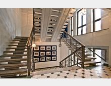 Treppenhaus mit mehreren Etagen und offenen Treppen aus Stein, flankiert von Geländern mit Metallstäben. Große Fenster an der Wand lassen viel Tageslicht einfallen, und an der Wand hängen gerahmte Schwarz-Weiß-Fotografien.