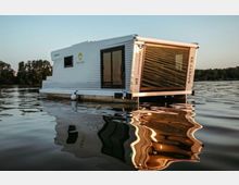 Ein modernes Hausboot mit weißen Metallwänden und großen Glasfenstern, das auf einem ruhigen Gewässer schwimmt, umgeben von Bäumen am Horizont bei Tageslicht.