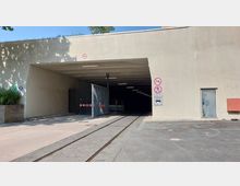 Einfahrt zu einem Tunnel mit Betonfassade, Schienen führen in den Tunnel hinein. An der rechten Seite befinden sich Schilder mit Verkehrs- und Zutrittsverboten sowie eine Metalltür, links ein kleines bepflanztes Steinbeet.