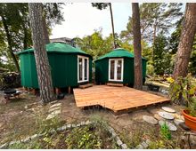 Zwei grüne, runde Zelte mit spitz zulaufenden Dächern stehen nebeneinander auf einem Waldgrundstück, umgeben von hohen Bäumen. Vor den Zelten liegt eine Holzterrasse, und im Vordergrund sind Gartenpflanzen sowie ein gepflasterter Weg sichtbar.