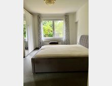 Helles Schlafzimmer mit großem Polsterbett und gepolstertem Kopfteil, grauem Teppichboden und einem Kleiderschrank mit Spiegeltüren. Ein breites Fenster mit hellen Vorhängen und Blick ins Grüne dominiert die Rückwand, darüber hängt eine Deckenleuchte.