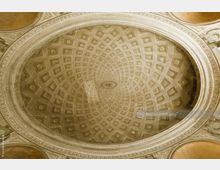 Gewölbte Decke eines historischen Gebäudes mit symmetrischem Kassettenmuster, verziert mit floralen Elementen in den Kassetten und ornamentalem Rahmen. Die Farben dominieren in Beige- und Goldtönen.