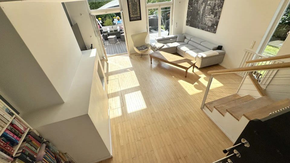 Heller, moderner Wohnbereich mit hellem Holzfußboden, großer L-förmiger Sofalandschaft, Couchtisch und hohen Fenstern mit Tür zur Terrasse und in den Garten. Rechts führt eine offene Treppe mit Geländer nach oben, an der Wand hängt ein großes Stadtbild.