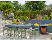 Gepflegter Garten mit einer Terrasse aus Steinfliesen, einer Sitzgruppe mit grünen Stühlen und einem Tisch aus Stein. Im Hintergrund ist eine Natursteinmauer, dichtes Grün, blühende Sträucher sowie eine Feuerschale zu sehen.