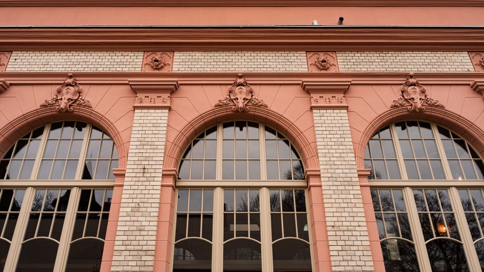 Fassade eines historischen Gebäudes mit drei großen Rundbogenfenstern, eingerahmt von rosafarbenen Stuckrahmen und weißen Ziegelpfeilern. Über den Fenstern befinden sich ornamentale Wappenreliefs und ein breites Gesims unter dem Dachrand.