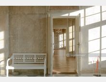 Heller, historischer Innenraum mit einer weißen Holzbank an einer Wand links und geöffneten Türen, die in einen mit Parkettboden ausgestatteten Raum mit Kronleuchter führen. Auf der rechten Seite steht ein Schirmständer mit einem roten Regenschirm, während Sonnenlicht durch große Fenster hereinfällt und Schatten auf den Boden wirft.