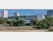 Städtische Wohnanlage mit mehreren Plattenbauten, umgeben von Bäumen und Grünflächen; im Vordergrund ein Zaun und eine unbefestigte Straße.