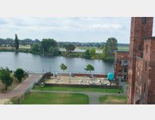 Blick auf einen Fluss mit einer Brücke im Hintergrund, umgeben von Bäumen und Wiesenlandschaft. Im Vordergrund ein kleiner Sandstrand mit Liegestühlen, Schirmen und Spielgeräten sowie ein rotes Backsteingebäude mit der Aufschrift "Strandbar & Café".