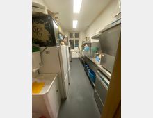 Schmaler, länglicher Küchen- und Spülraum mit grauem Boden, Deckenleuchten und einem Fenster am Ende. Links stehen Kühlschränke und Regale, rechts verläuft eine Edelstahl-Arbeitszeile mit Spülbecken, Abtropffläche und großen Küchengeräten.