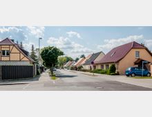 Eine ruhige Wohnstraße mit Einfamilienhäusern, von denen einige Fachwerk- oder Satteldächer haben. Die Straße ist gesäumt von Bäumen und Gehwegen, mit parkenden Autos auf beiden Seiten.