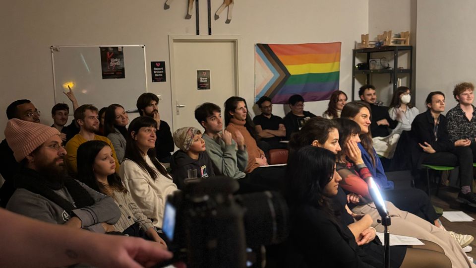 Indoor event space with rows of chairs facing a small presentation area, set in a white-walled room with exposed ceiling beams and track lighting. Wall decor includes a mounted antelope sculpture above a door, several posters, and a large pride flag hanging beside metal shelving.
