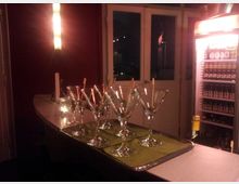 Eine Bar mit einem geschwungenen Tresen, auf dem mehrere Martini-Gläser auf einem Tablett stehen. Im Hintergrund sind eine beleuchtete Wand, eine Glastür mit nächtlichem Ausblick und ein beleuchteter Getränkekühlschrank mit Flaschen sichtbar.