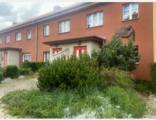 Mehrfamilienhaus mit rötlicher Fassade und weißen Fensterrahmen, teilweise mit Büschen und Blumen bepflanztem Vorgarten. An der Wand befindet sich ein Relief und eine Tafel, umgeben von Grünpflanzen.