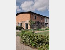 Backsteingebäude mit Elementen aus Feldsteinen, umgeben von einer gepflegten Hecke, Blumenbeeten und Grünflächen. Der Himmel ist leicht bewölkt, und ein Ziegelweg führt entlang des Gebäudes.