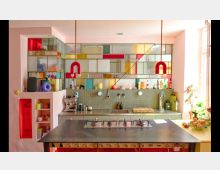 Moderne Küche mit bunten, geometrischen Glasregalen an der Wand, grauer Arbeitsfläche mit integriertem Gasherd, zwei roten Hängeleuchten und hellen rosa Wänden. Rechts ein Fenster, das den Raum mit natürlichem Licht durchflutet, und Pflanzen sowie Küchenutensilien auf den Arbeitsflächen.