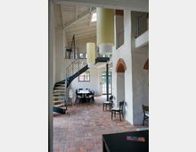 Innenraum eines Gebäudes mit hell verputzten Wänden und freiliegender Holzbalkendecke. In der Mitte steht eine Wendeltreppe aus Metall, umgeben von Tischen und Stühlen, auf einem Boden aus roten Ziegelsteinen.