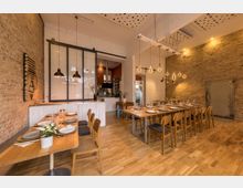 Gemütlicher Innenraum eines Restaurants mit sichtbaren Ziegelwänden und Holzböden. Die Tische sind gedeckt, Hängelampen mit verschiedenen Leuchtendesigns beleuchten den Raum, und im Hintergrund ist eine offene Küche mit Glasabtrennung sichtbar.