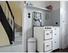 Kleine Küche mit Retro-Ofen in Weiß, daneben weiße Schränke und eine graue Rückwand mit hängenden Küchenutensilien. Links führt eine Treppe mit schwarzem Handlauf nach oben.