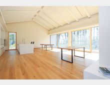 Modernes Wohnzimmer mit offenem Grundriss, hellem Holzfußboden und einer weißen Decke mit sichtbaren Balken. Große Fenster mit Holzrahmen lassen viel Tageslicht herein und bieten Ausblick auf eine Baumlandschaft; in der Mitte stehen ein Esstisch und eine Küche mit Insel.