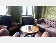 Ein gemütliches Wohnzimmer mit klassischen Sitzmöbeln, darunter ein brauner Sessel und ein lila Chesterfield-Sofa. Im Mittelpunkt steht ein runder Holztisch, auf dem Gläser, eine Fernbedienung und ein Behälter stehen; im Hintergrund sind eine grüne Tapete mit Blumenmuster, ein Fenster mit Vorhängen und ein Heizkörper sichtbar.