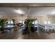 Industriell gestalteter Veranstaltungsraum mit Betonwänden, hoher Decke und Fenstern im Bogenstil. Links befinden sich Sitzecken mit schwarzen Sofas, Tischen und Pflanzen, während rechts Reihen weißer Stühle aufgestellt sind.