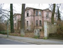 Ehemaliges zweistöckiges rosa Gebäude in einem verwahrlosten Zustand, mit zerstörten Fenstern und einer beschädigten Fassade, umgeben von kahlen Bäumen und Gestrüpp. Im Vordergrund befinden sich eine kleine Einfahrt mit beschädigter Steinmauer und eine Straße mit Gehweg.