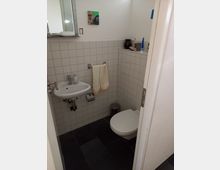 Kleines Badezimmer mit weiß gefliesten Wänden und dunklem Fliesenboden. Zu sehen sind eine Wandtoilette, ein kleines Waschbecken mit Handtuchhalter sowie ein Spiegel und Ablageflächen mit Alltagsgegenständen.