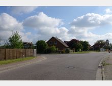 Straßenszene in einem Wohngebiet mit Einfamilienhäusern, die von Gärten und Holzzäunen umgeben sind. Am Straßenrand befindet sich ein Gehweg, und im Hintergrund ist ein bewölkter Himmel mit einzelnen blauen Lücken zu sehen.