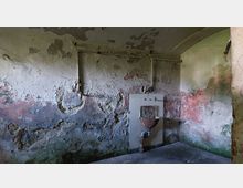 Ein verlassener Raum mit einer stark verfärbten, bröckelnden Wand und sichtbaren Rohren. Im Raum befindet sich ein alter, rostiger Waschbeckenaufsatz an einer Kachelfläche, umgeben von abblätterndem Putz und Wasserflecken.
