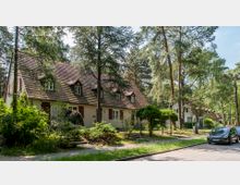 Eine von Bäumen umgebene Wohnstraße mit mehreren Reihenhäusern im traditionellen Stil, gekennzeichnet durch Satteldächer mit kleinen Gauben und hell getünchte Fassaden. Im Vordergrund sind ein Gehweg, Bepflanzungen und ein geparktes Auto zu sehen.