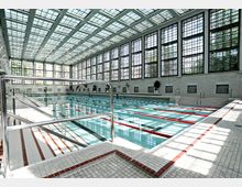 Innenansicht eines großen, lichtdurchfluteten Schwimmbads mit hohen, gläsernen Fensterfronten und einem rechteckigen Becken mit abgetrennten Bahnen. Die Architektur ist geprägt von klaren Linien, weiß gefliesten Böden und roten Markierungen entlang der Beckenumrandung.