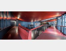 Moderne Innenansicht eines Gebäudes mit roten Rampen und einem geschwungenen roten Deckenelement. Die Wände sind dunkelgrau, und große Fensterfronten auf der rechten Seite lassen Tageslicht herein; im hinteren Bereich sind weitere offene Bereiche und Sitzgelegenheiten erkennbar.