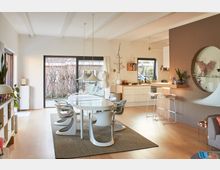 Helles modernes Esszimmer und Küche mit offener Bauweise, weißen Möbeln und Holzfußboden. Große Fenster mit Blick auf den Garten, ein ovaler Esstisch mit Designerstühlen und eine Küchenzeile mit Arbeitsfläche dominieren den Raum.