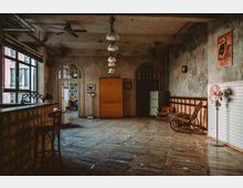 Großer, rustikaler Innenraum mit einer hohen Decke, freiliegenden Rohren und abgenutzten Wänden. Der Raum enthält Holzmöbel, eine Theke, eine Waage sowie einen Schaukelstuhl, mit einer Mischung aus vintage Dekor und industriellem Stil, und großen Fenstern, die Tageslicht hereinlassen.