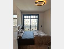 Modernes Schlafzimmer mit Doppelbett und gepolstertem Kopfteil, schlichtem Interieur und ringförmiger Deckenleuchte. Große schwarze Glas-Schiebetür führt auf einen Balkon mit Blick auf umliegende Wohngebäude.