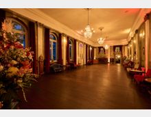 Historischer Ballsaal mit hohen Decken, verzierten Kronleuchtern und großen Rundbogenfenstern, umrahmt von schweren roten Vorhängen. Der Raum ist mit antiken Möbeln, wie Sofas und Sesseln, sowie Blumenarrangements dekoriert und in warmes, gelbes Licht getaucht.